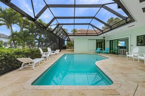 A swimming pool with a glass ceiling, surrounded by white lounge chairs, and a view of palm trees and houses. - Fully Remodeled Single-Story Home w/Heated Pool and Boat Dock (Marco Island)