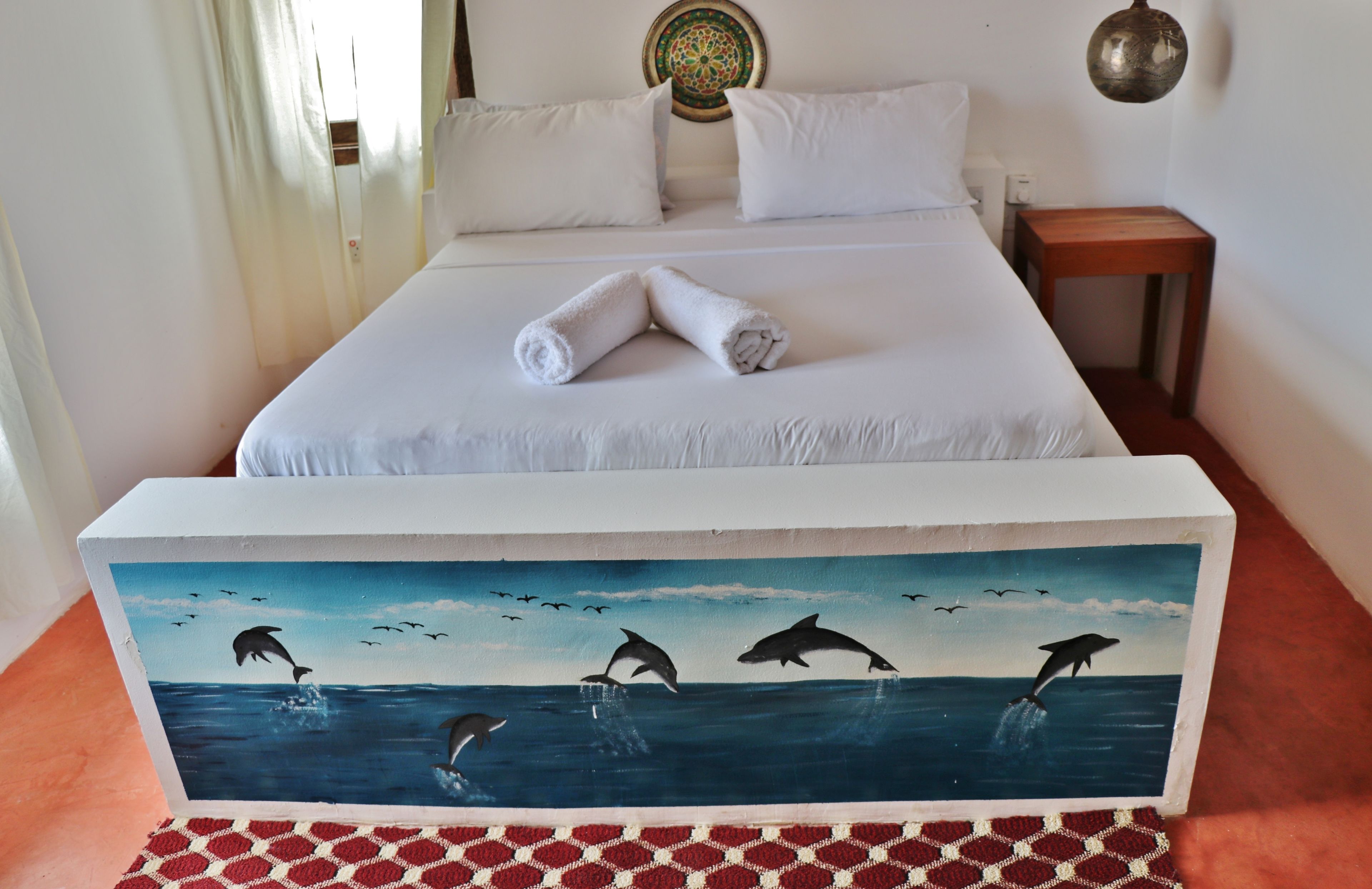 A hotel room with a bed, a nightstand, and a painting of dolphins on the floor.