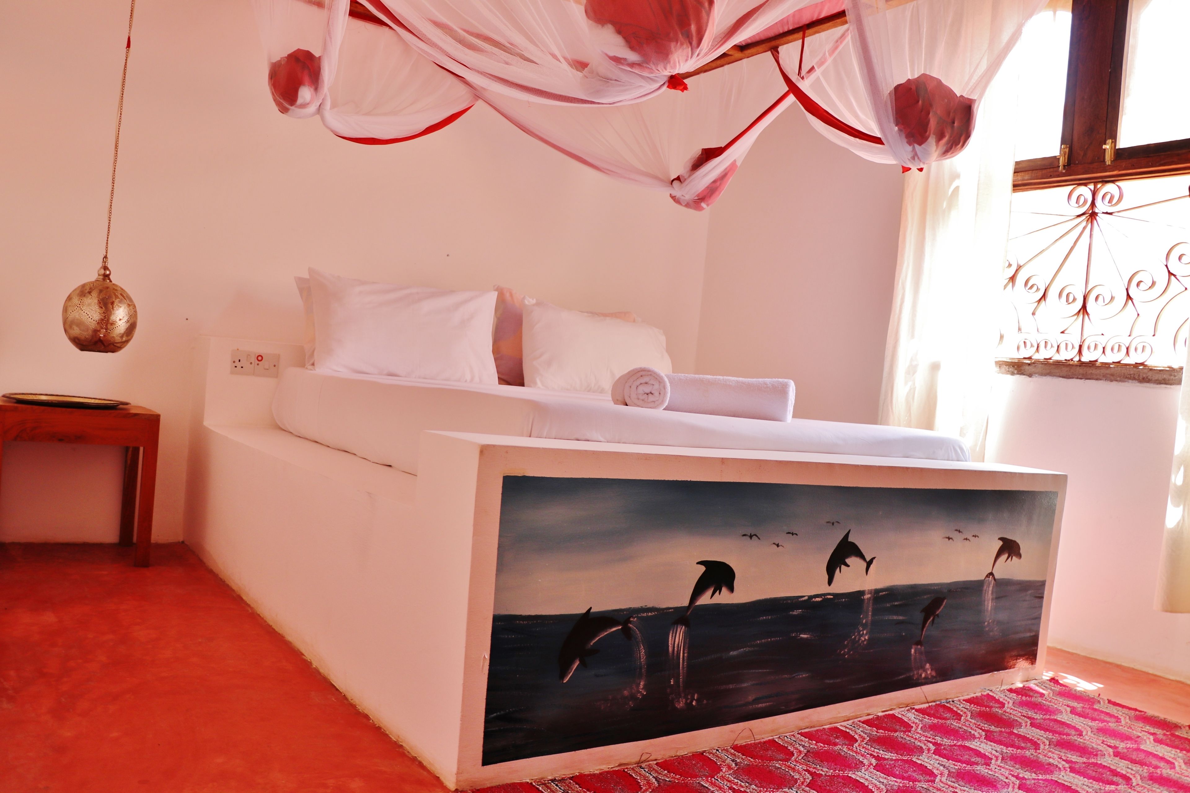 Un dormitorio con cama, una mesita de noche, una lámpara colgante y una ventana con cortinas.