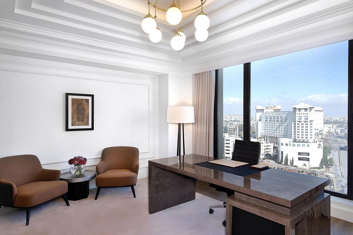 A modern hotel room with a large window offering a city view, a desk with a chair, two armchairs, and a floor lamp.