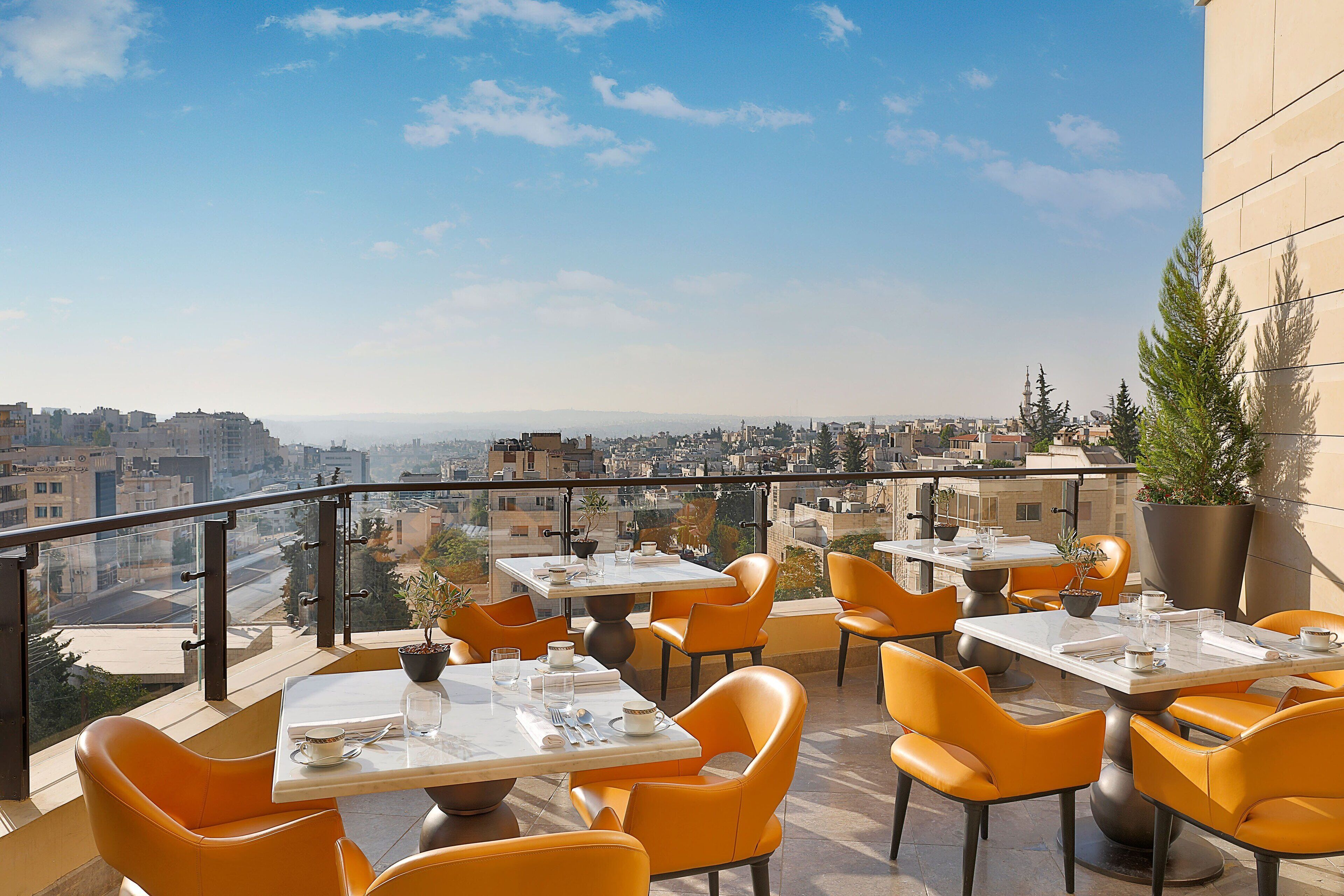 A rooftop terrace with orange chairs and tables, offering a city view.