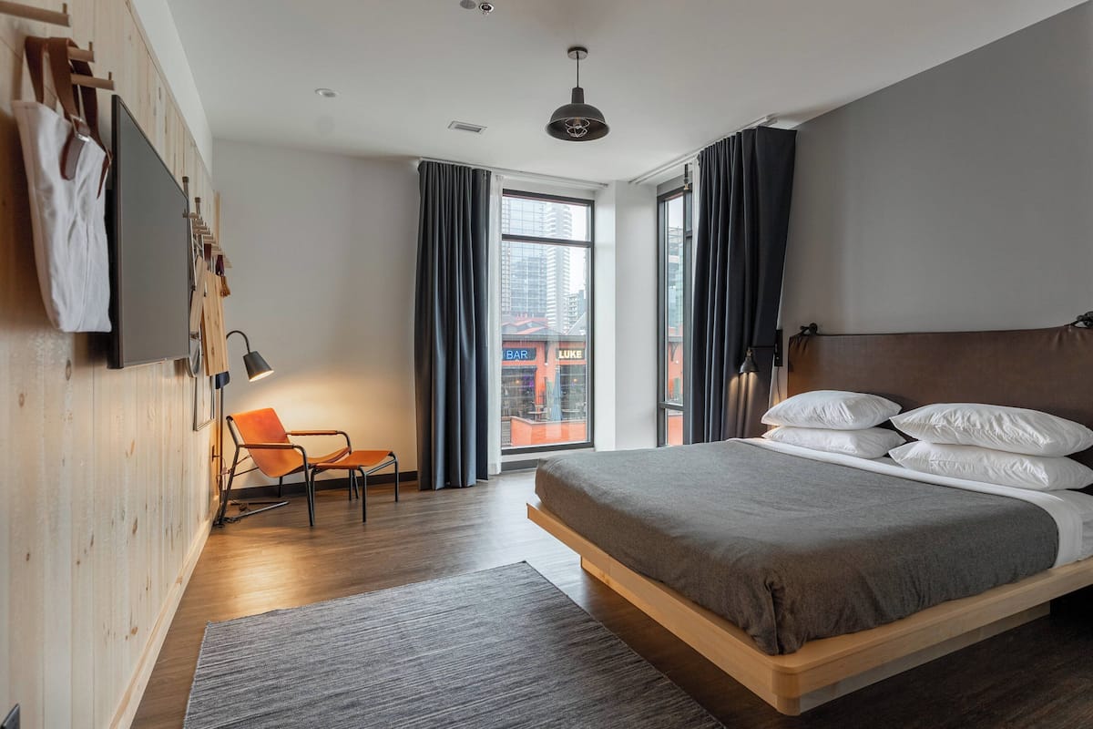 A modern bedroom with a wooden bed, a chair, a desk, and a large window with curtains.