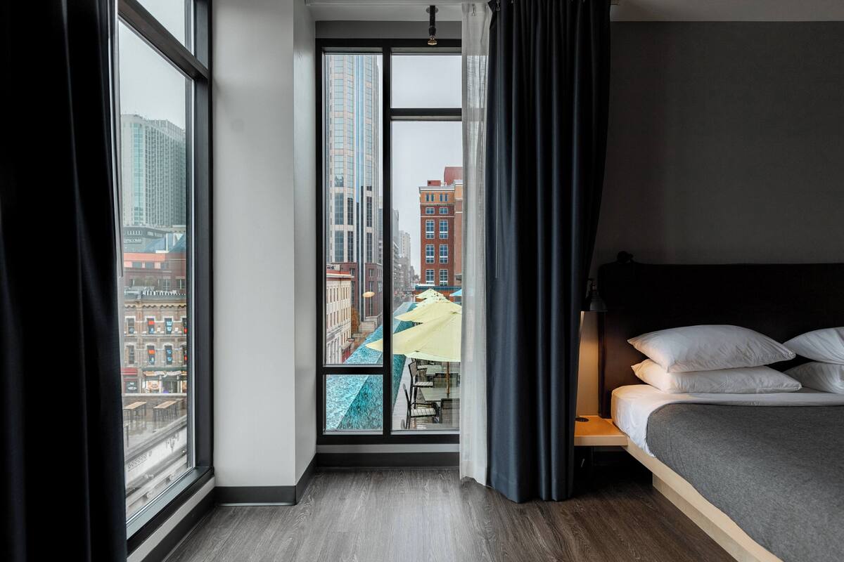 A hotel room with a large window, a bed, and a view of the cityscape.