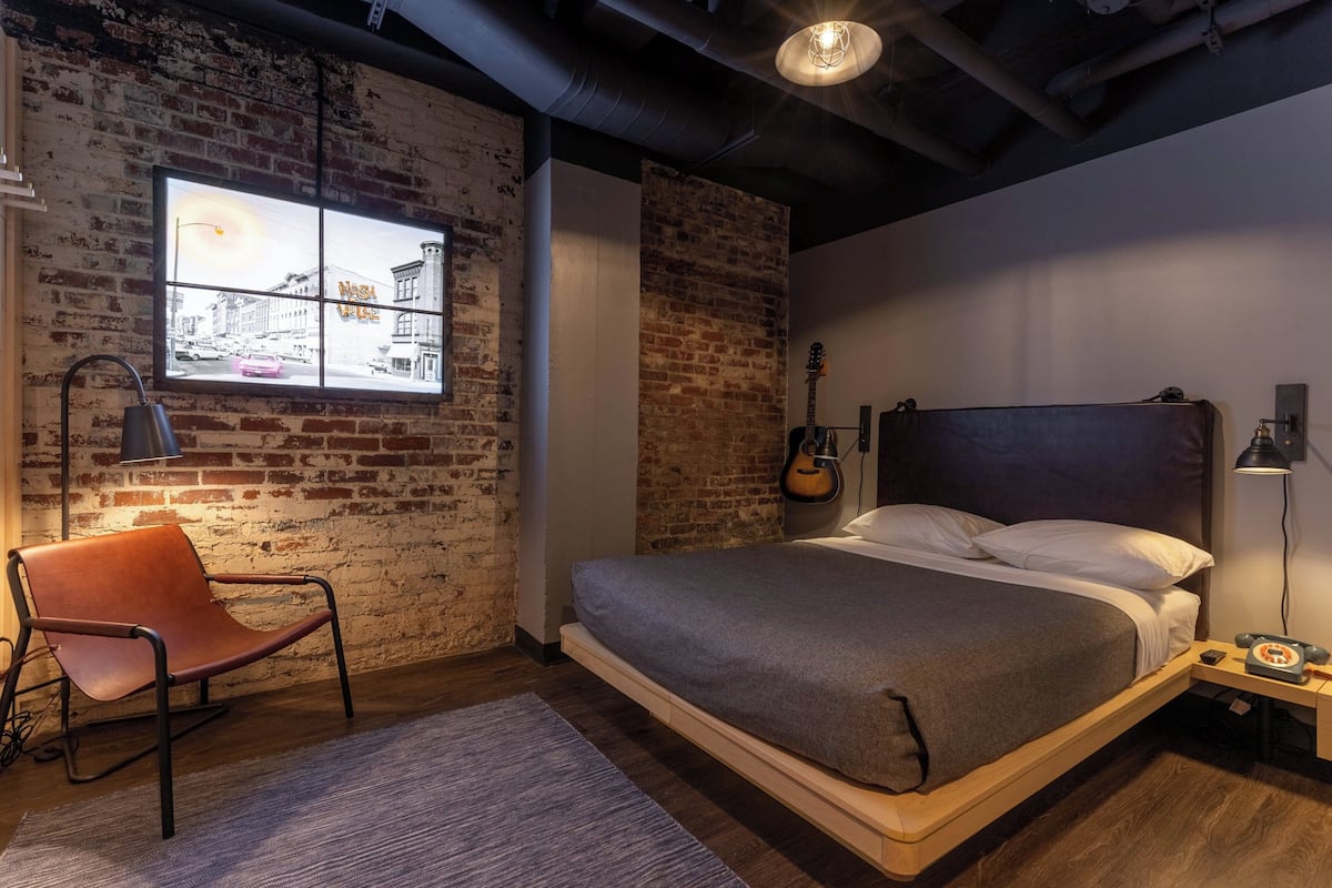 A bedroom with a loft bed, a brick wall, a window with a cityscape view, a guitar, and a vintage telephone.