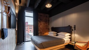 A modern hotel room with a large bed, a wooden headboard, a desk with a chair, a window with curtains, and a brick wall.