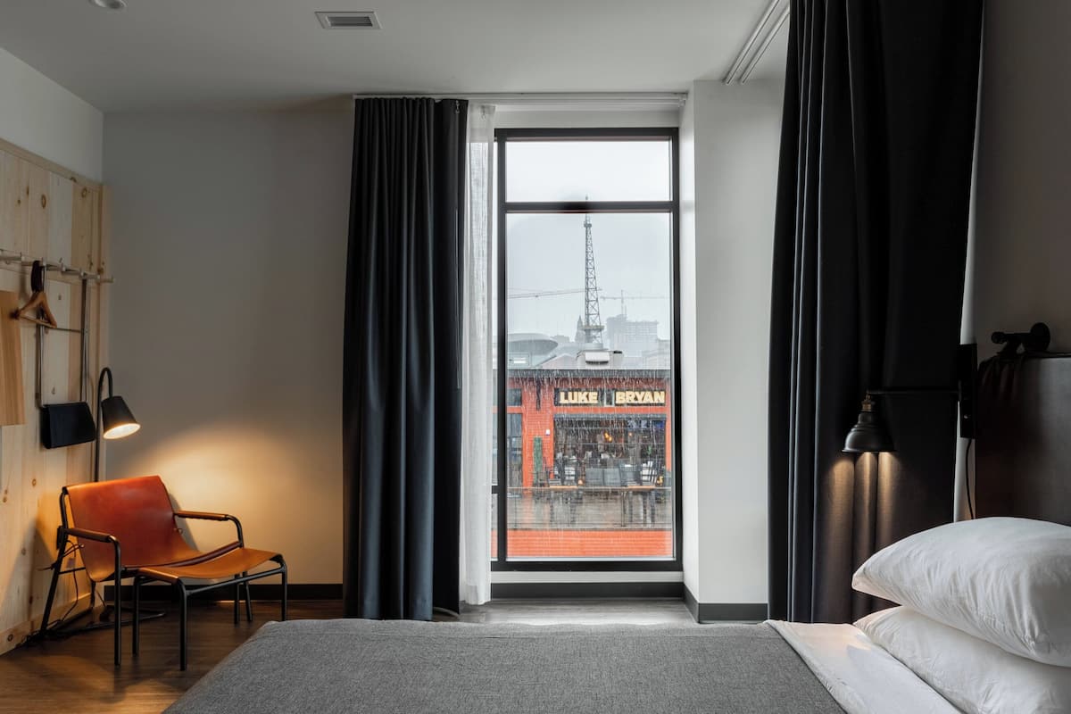 A modern hotel room with a large window, a bed, a chair, and a lamp.