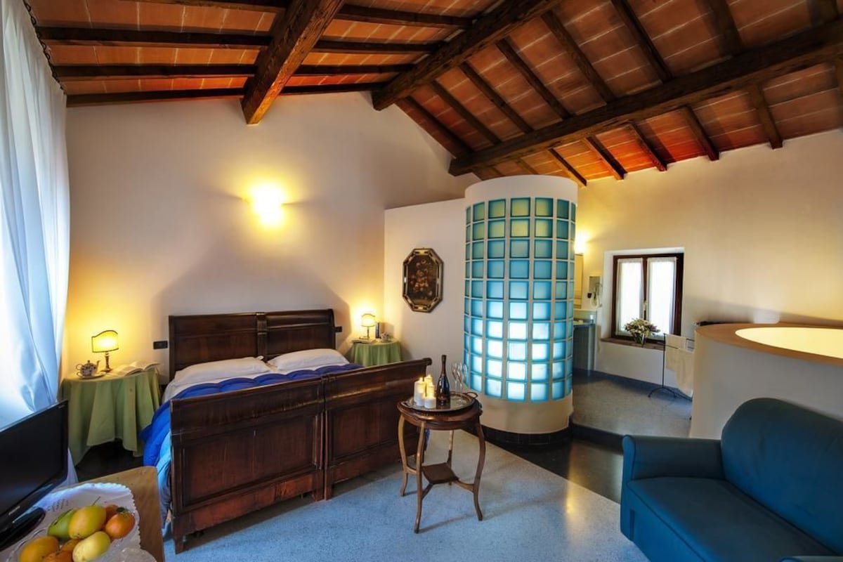 A bedroom with a wooden bed, a nightstand with a lamp, a blue cylindrical light fixture, and a window with a view.