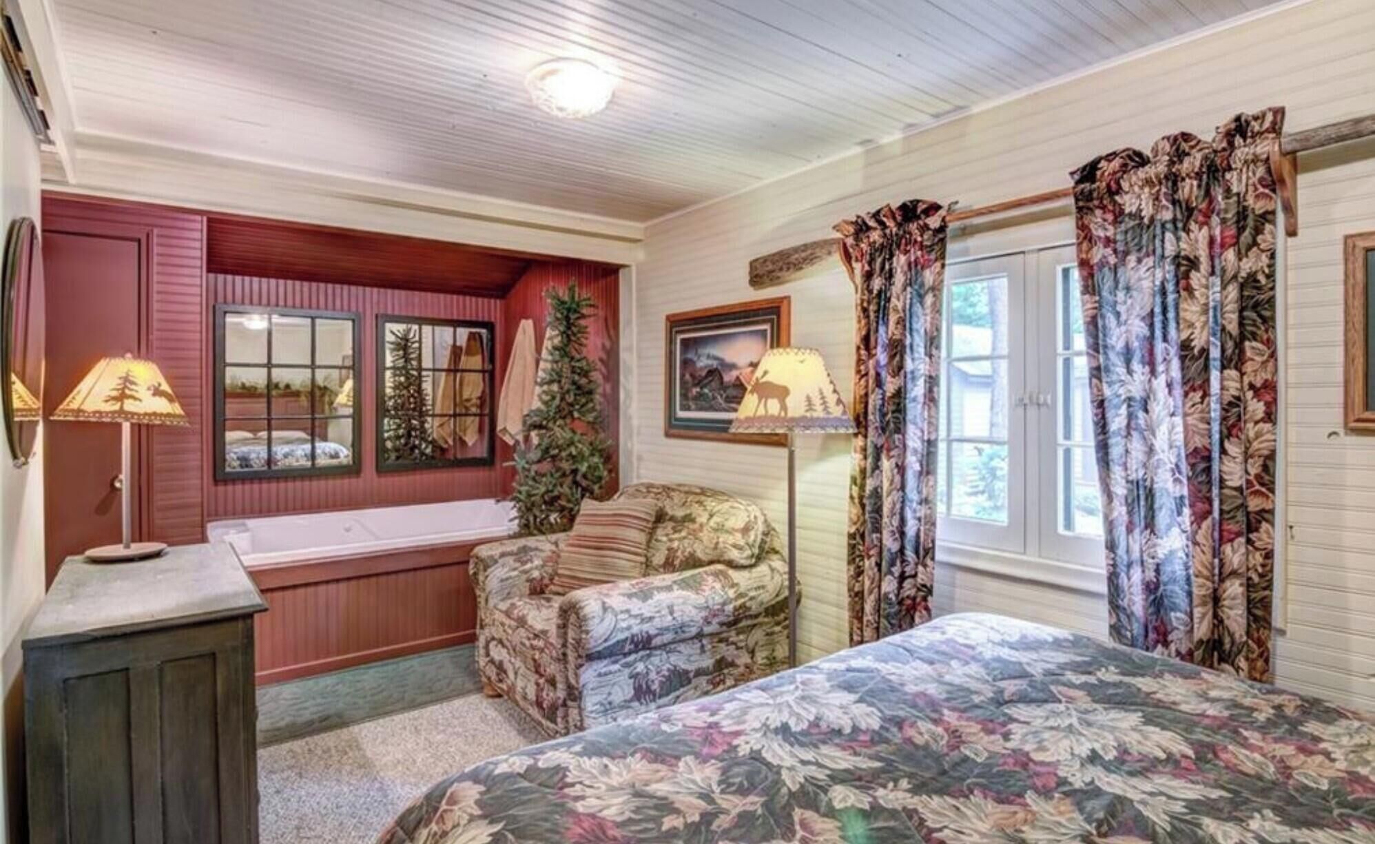 A cozy room with a bed, a chair, a window with curtains, a lamp, and a wall-mounted picture.