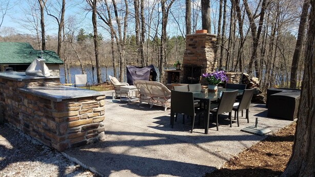 An outdoor patio with stone walls, a stone fireplace, wicker furniture, and a view of a lake.