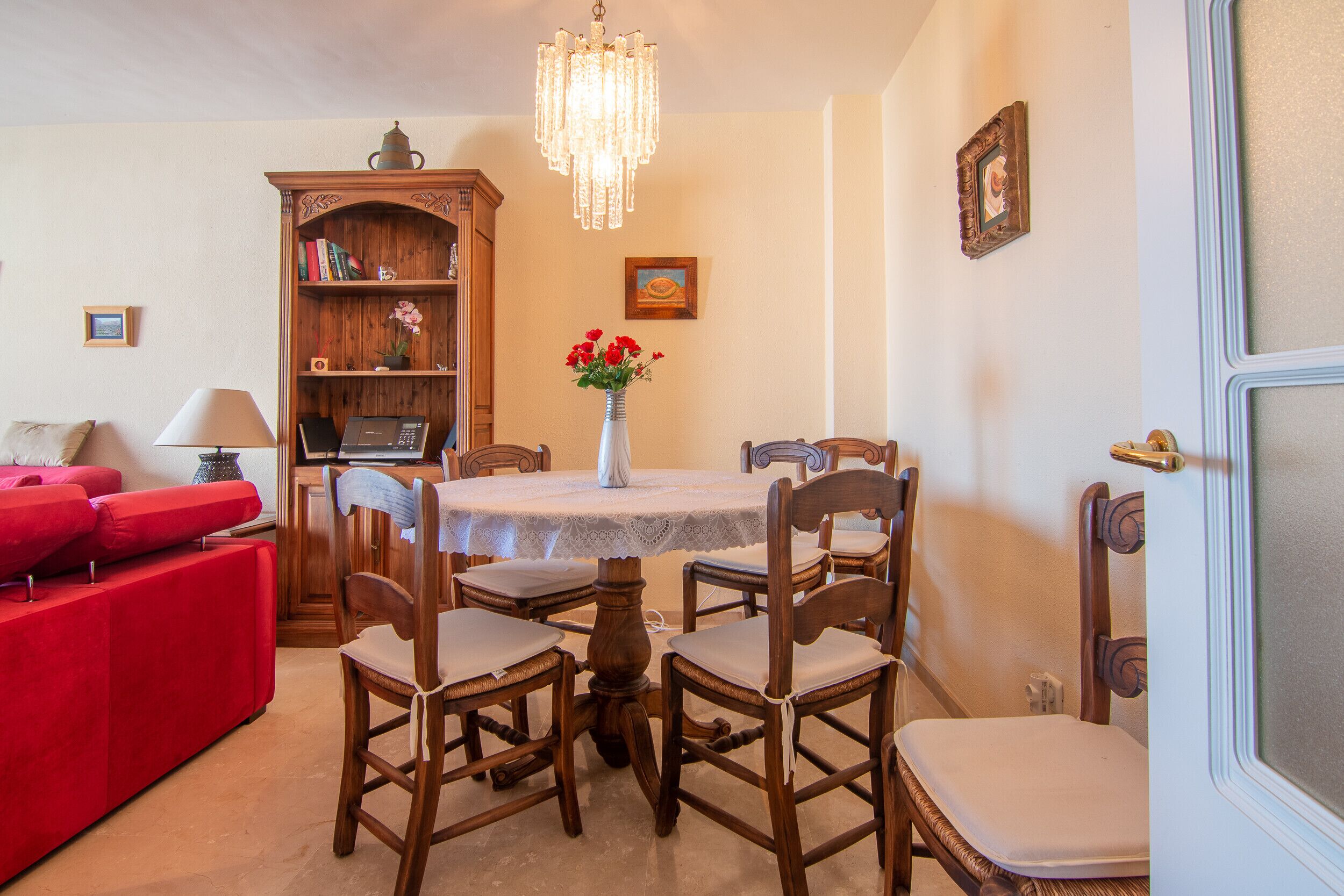 A dining area with a round table, six wooden chairs, a chandelier, a bookshelf, and a red sofa.