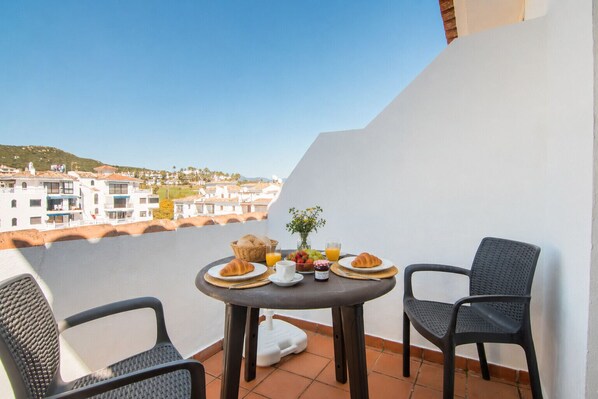 A balcony with a table set for breakfast, two chairs, and a view of buildings. - Marina Real Penthouse 2233 (Manilva)