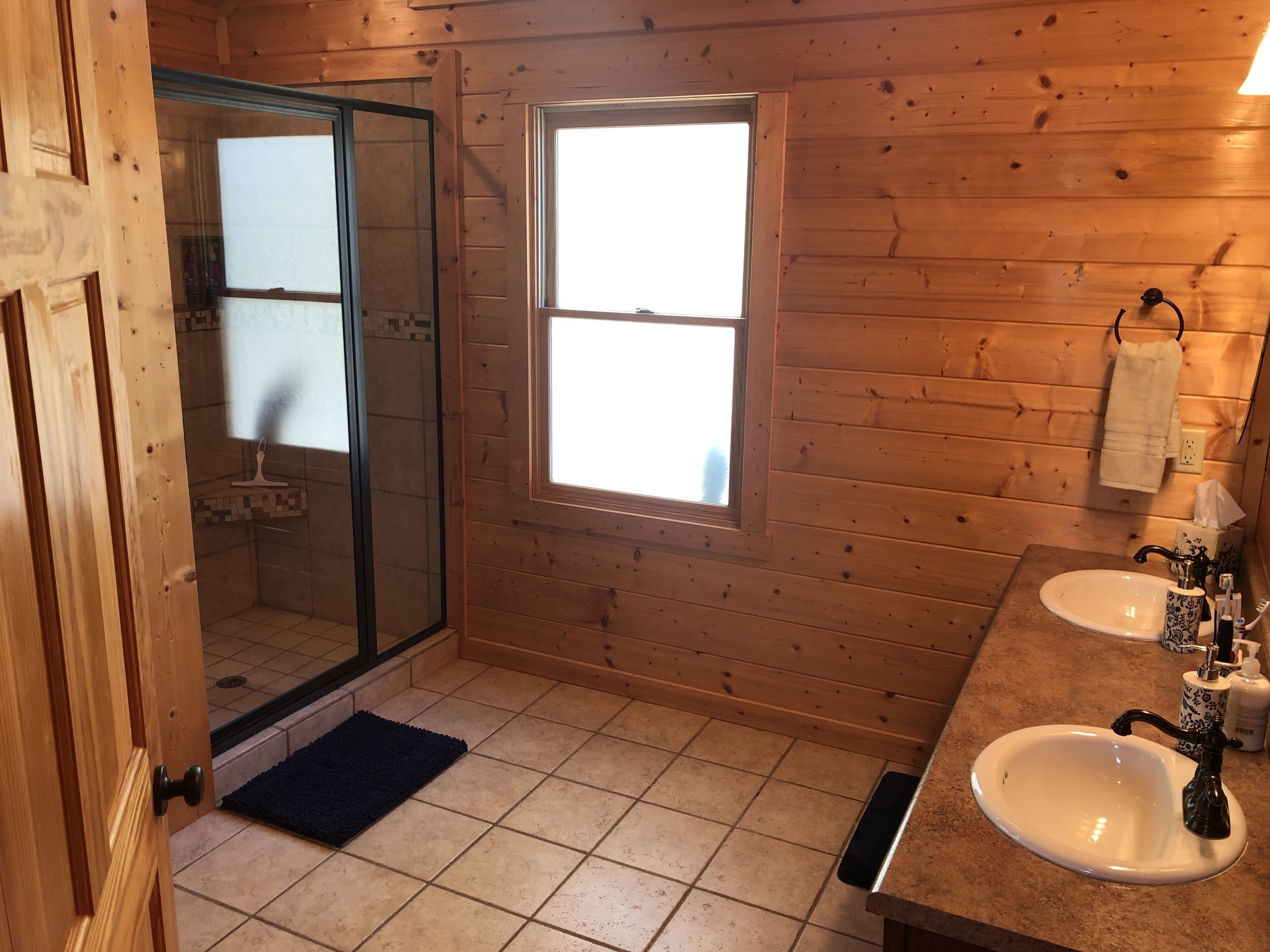 A wooden cabin bathroom with a glass shower door, a window, two sinks, and a towel hanging on the wall.