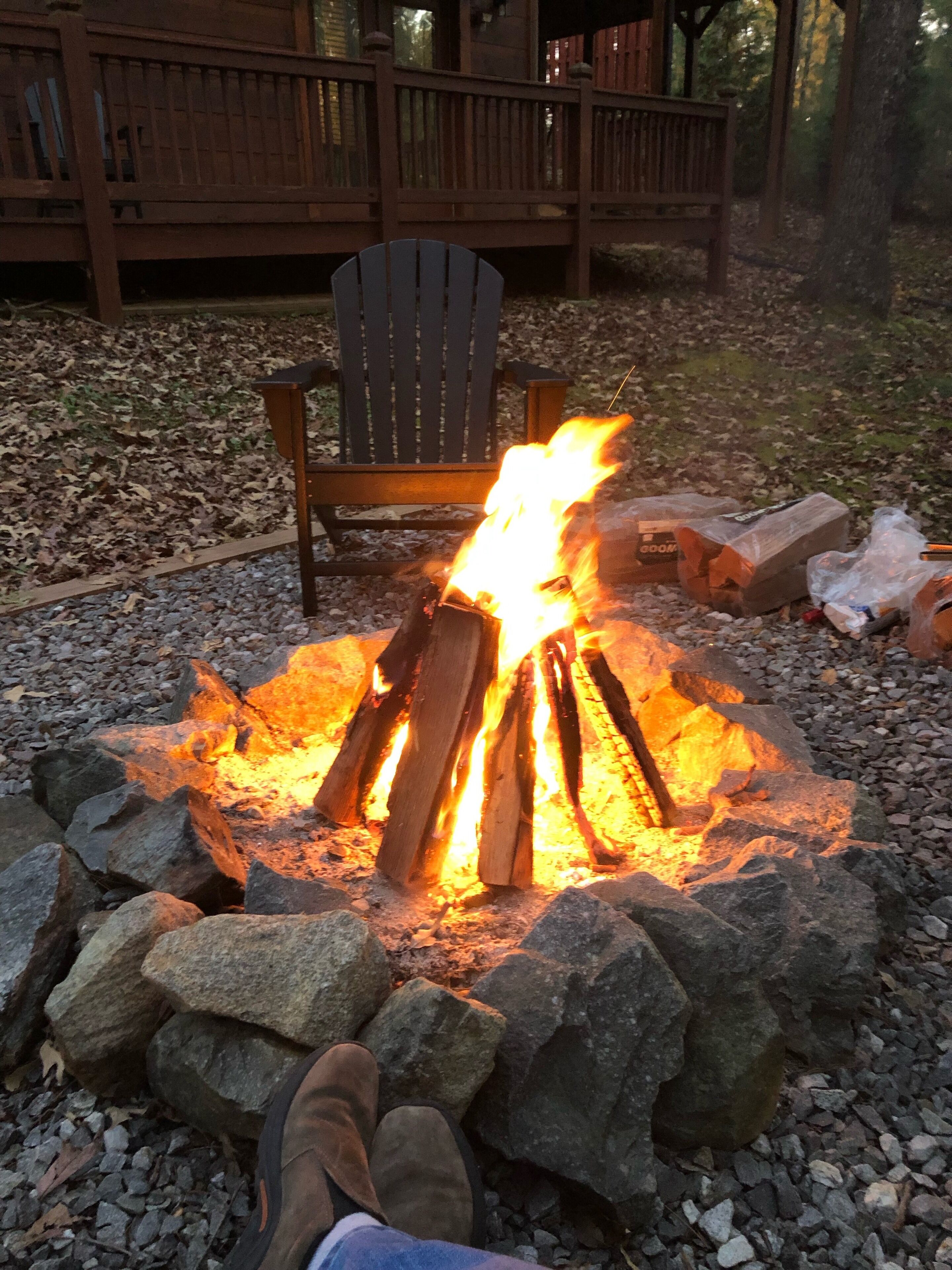 A campfire with logs and rocks, a wooden chair, and a deck in the background.