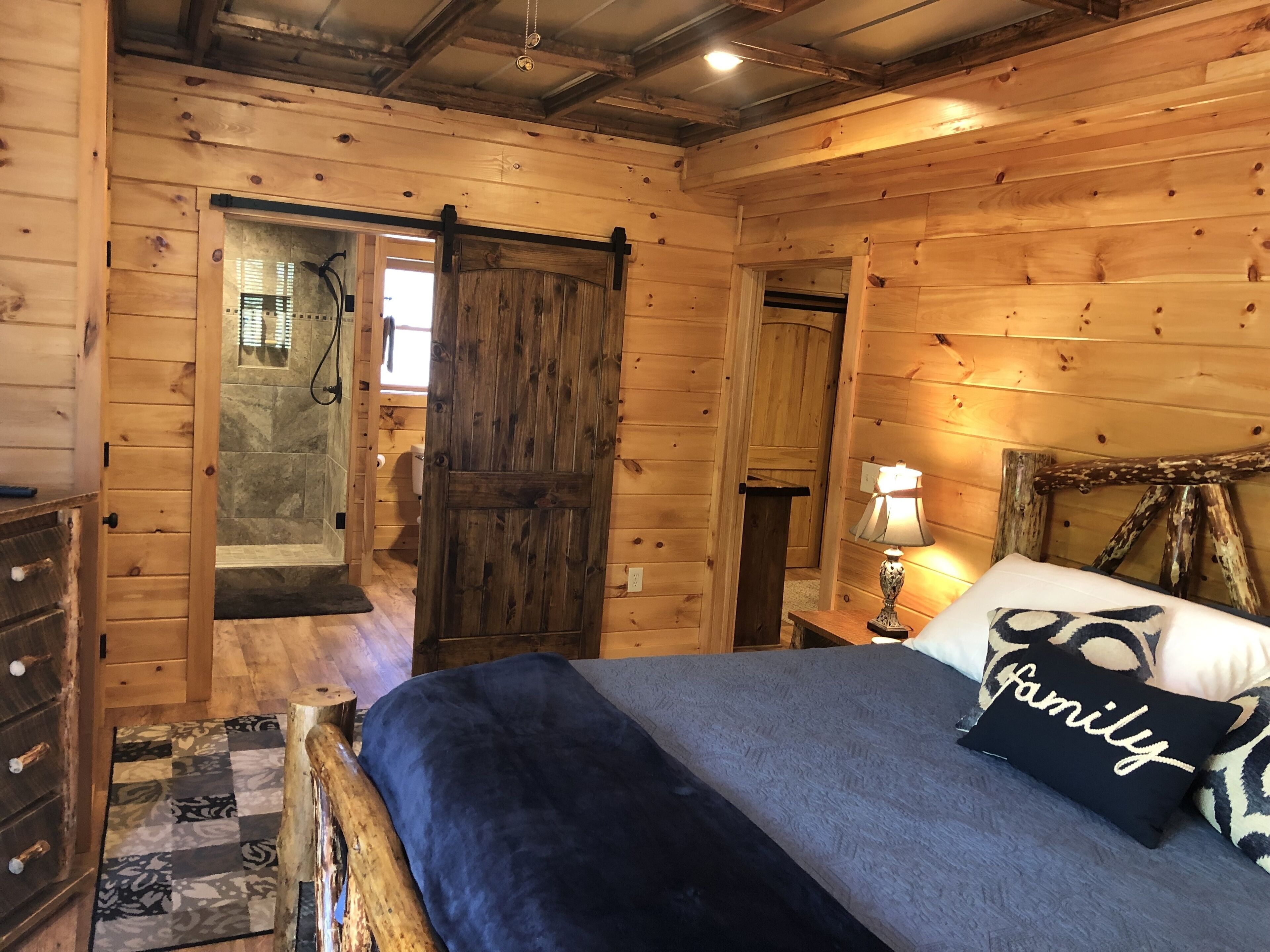 Quarto de cabana de madeira com uma cama, mesa de cabeceira, abajur e uma porta que dá para outro cômodo.