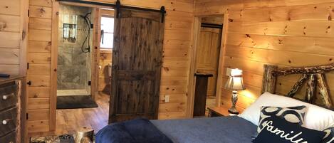 Quarto de cabana de madeira com uma cama, mesa de cabeceira, abajur e uma porta que dá para outro cômodo.