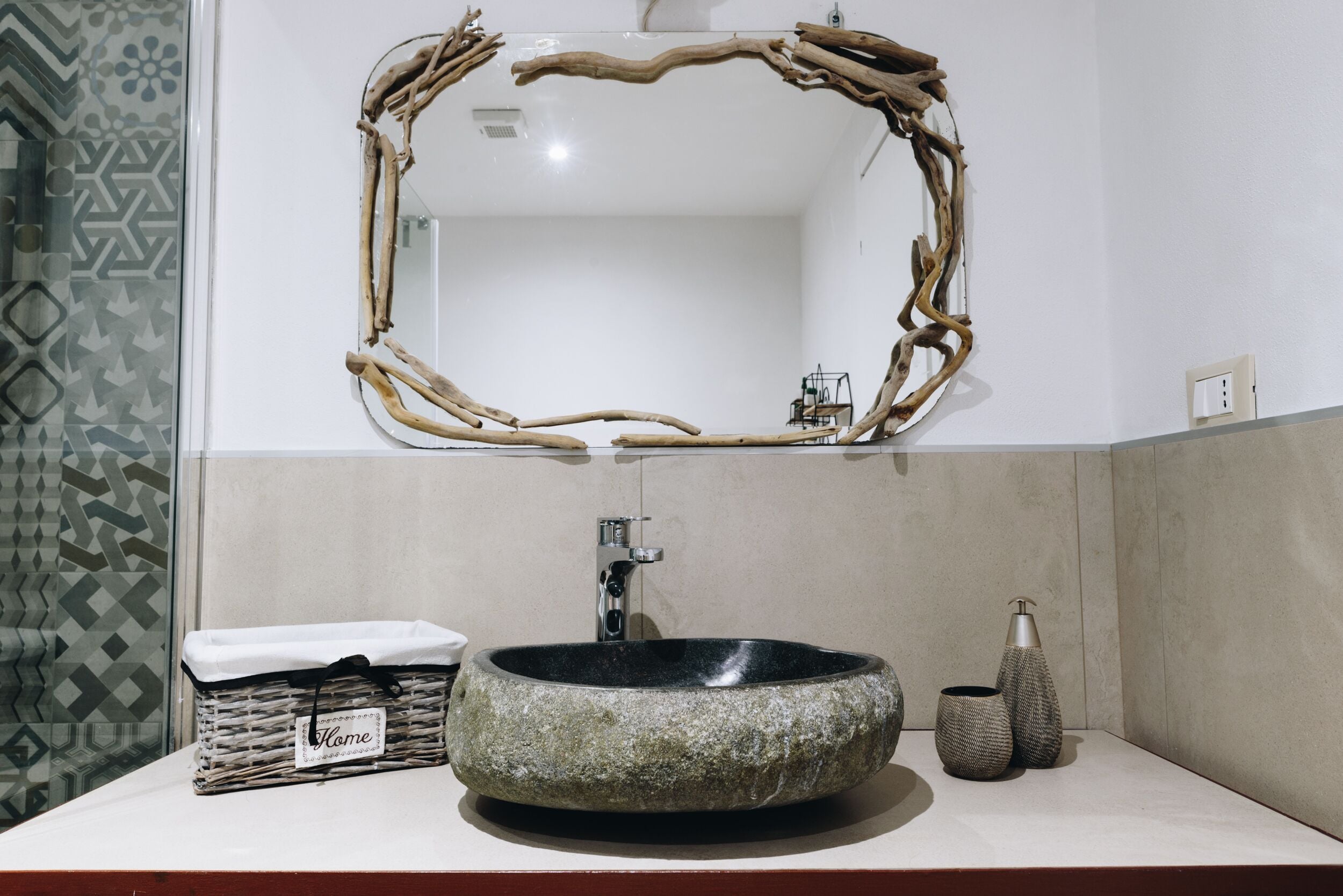 Une salle de bain avec un lavabo en pierre, un miroir encadré de branches, un panier portant l’inscription « Chez soi » et deux vases à motifs.