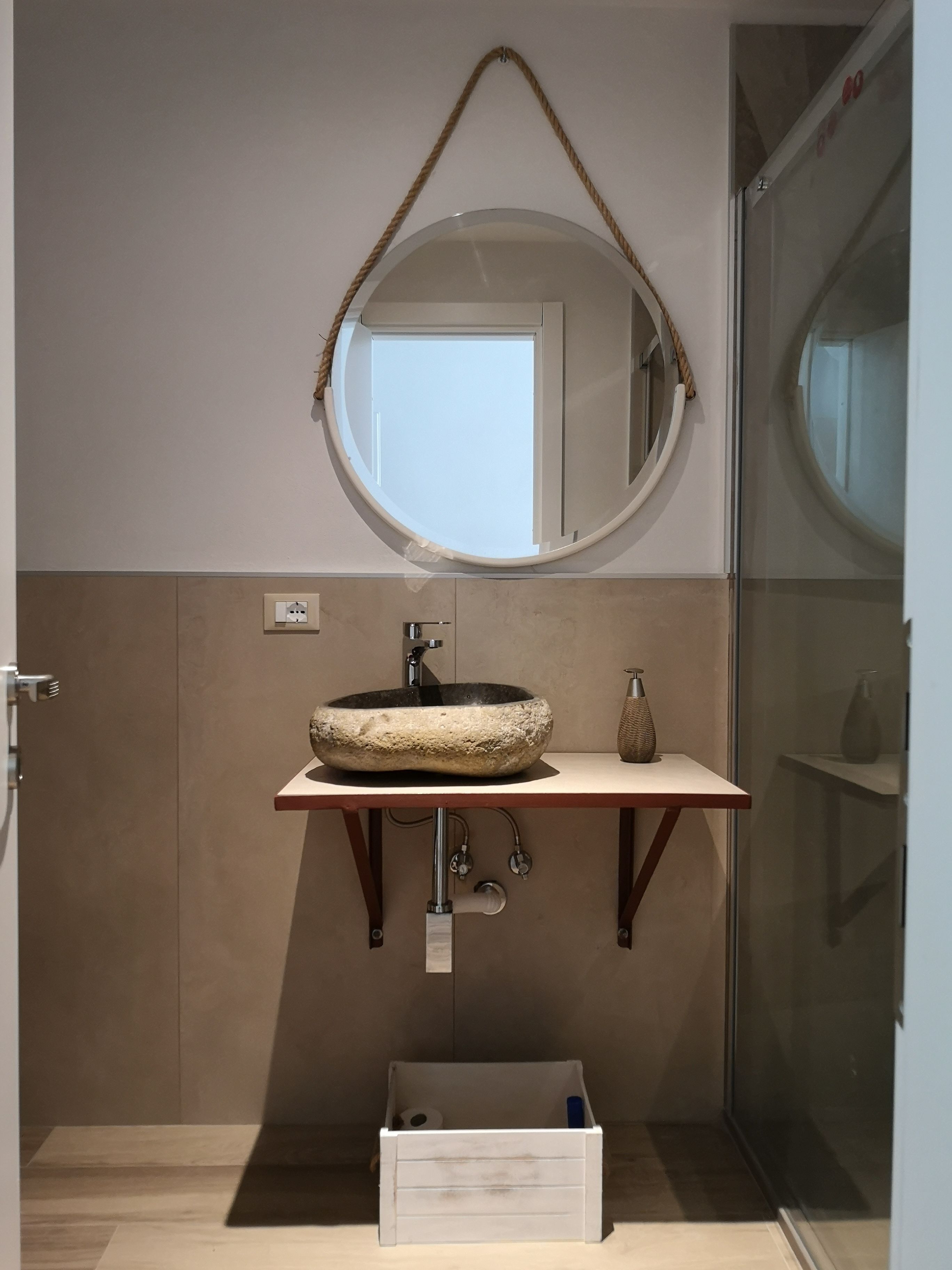 A bathroom with a round mirror, a stone sink, and a small vase on a shelf.