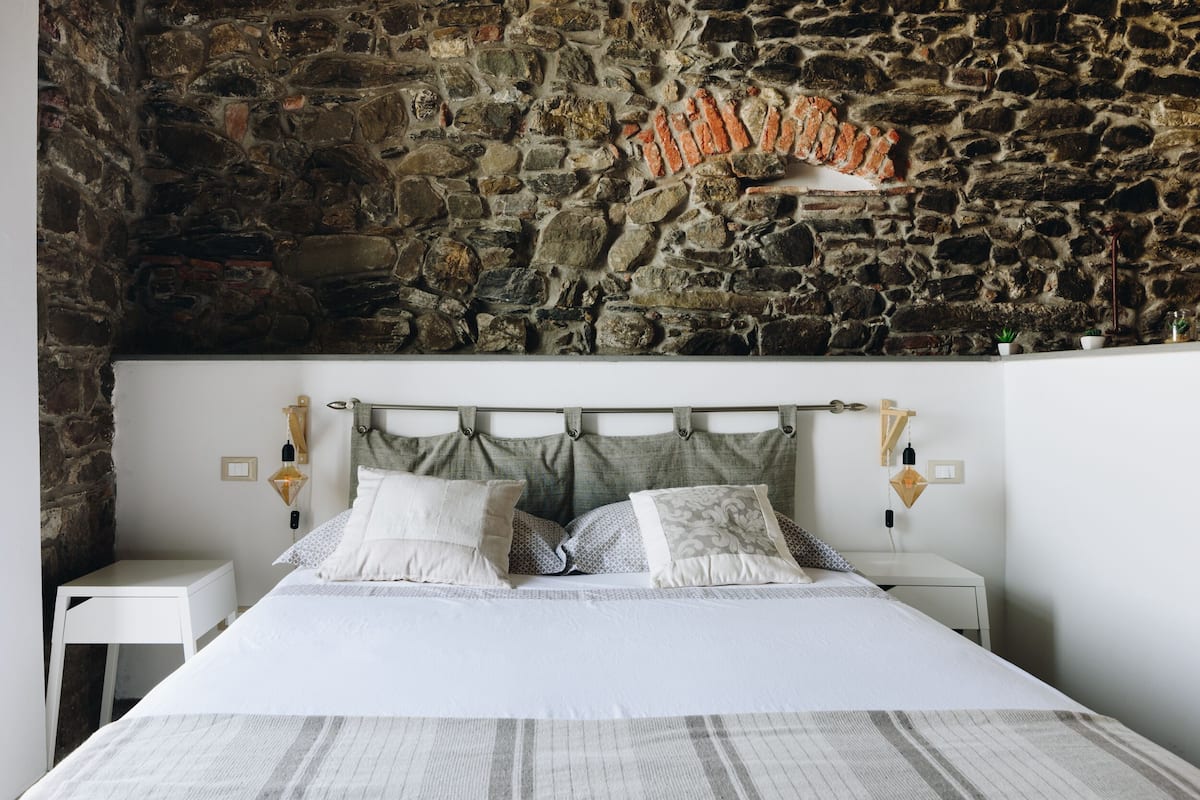 Un dormitorio con pared de piedra, una cama con ropa de cama blanca, dos mesitas de noche y lámparas fijadas a la pared.