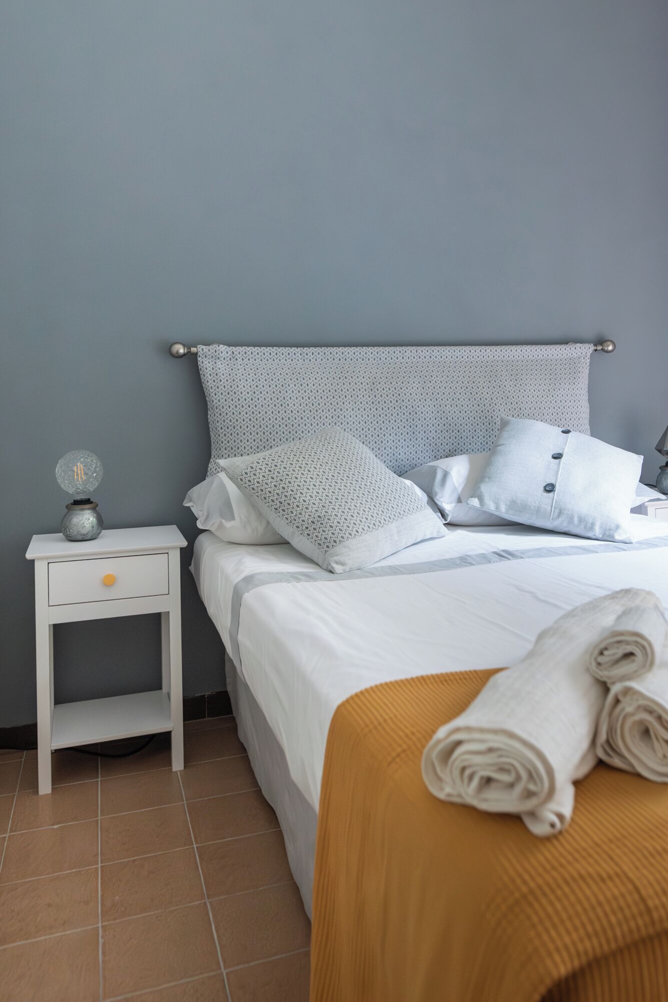 Un letto rifatto con biancheria bianca e diversi cuscini, un comodino con una lampada e un asciugamano arrotolato sul pavimento.