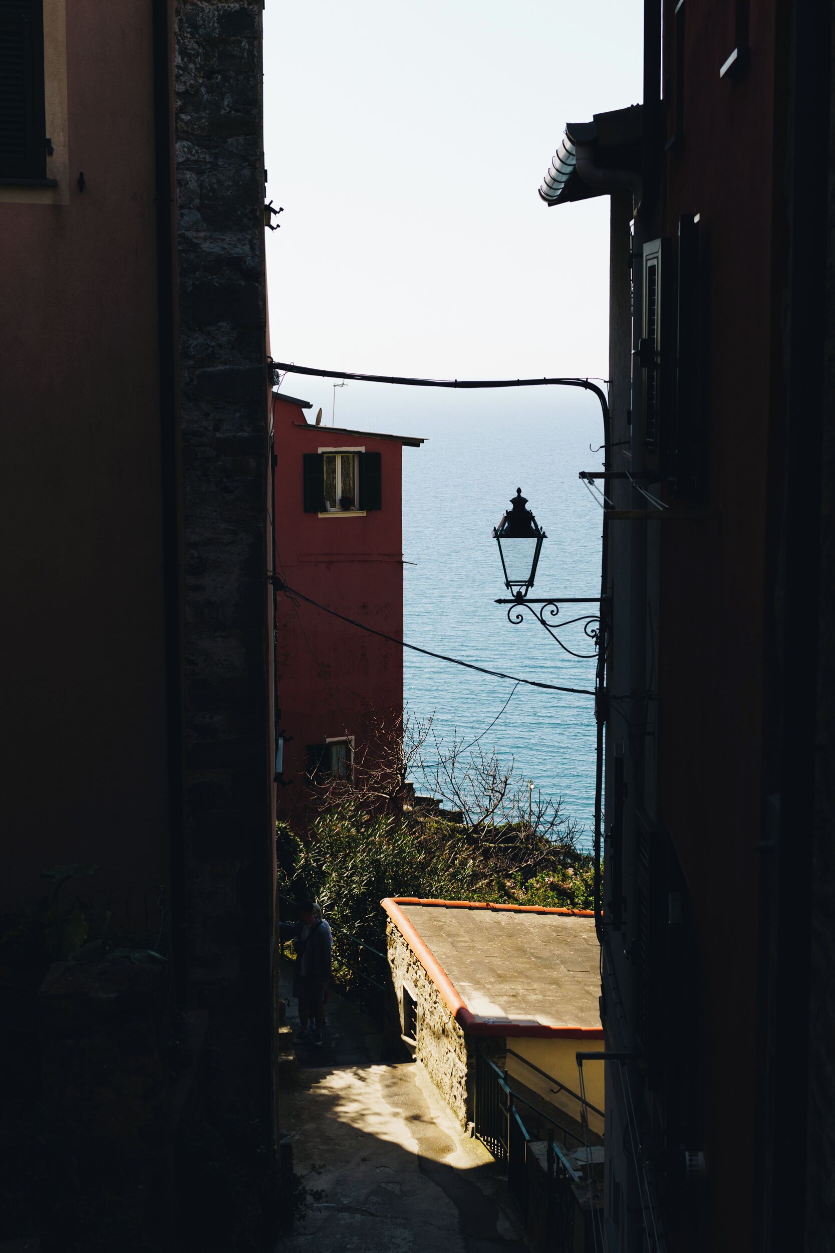 A narrow alleyway with a red building, a street lamp, and a view of the sea.