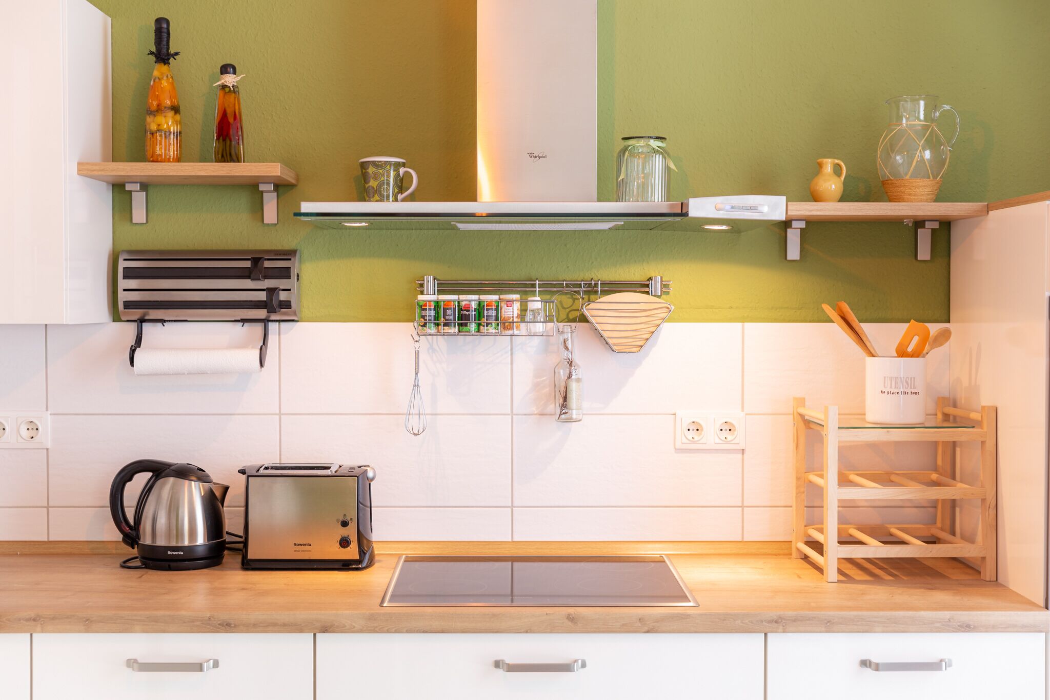 Une cuisine moderne avec des meubles blancs, une bouilloire en acier inoxydable, un grille-pain et un présentoir en bois contenant des ustensiles de cuisine.