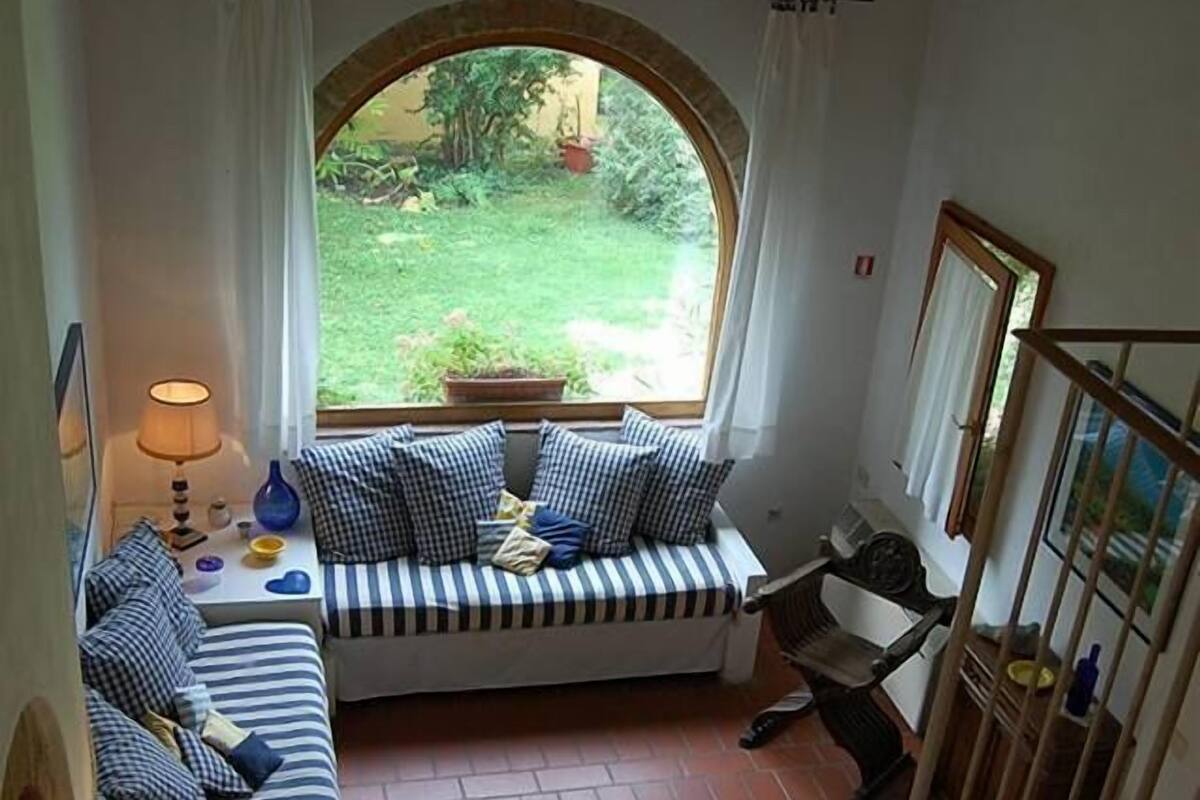 Una habitación acogedora con un ventanal curvo, un sofá con cojines a rayas, un sillón amarillo y una mesita con lámpara.
