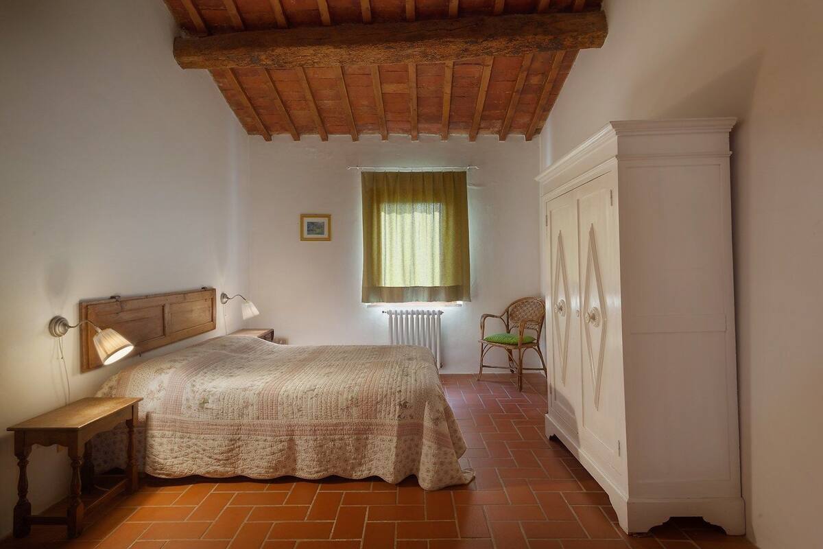 Un dormitorio con cabecera de madera, cama con colcha estampada, mesita de noche de madera, silla de mimbre, radiador y armario blanco.