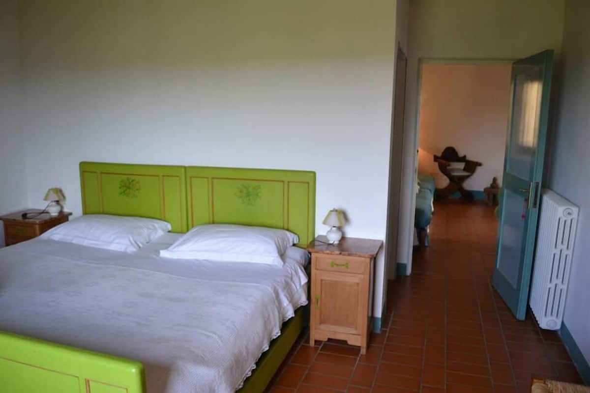 Un dormitorio con una cama verde, dos mesitas de noche con lámparas, una mesita auxiliar de madera y un radiador.