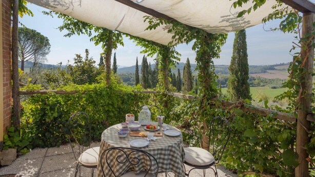Un espace de restauration en plein air avec une table dressée pour quatre personnes, entouré d’une végétation luxuriante et offrant une vue pittoresque sur des collines ondulantes et des arbres.