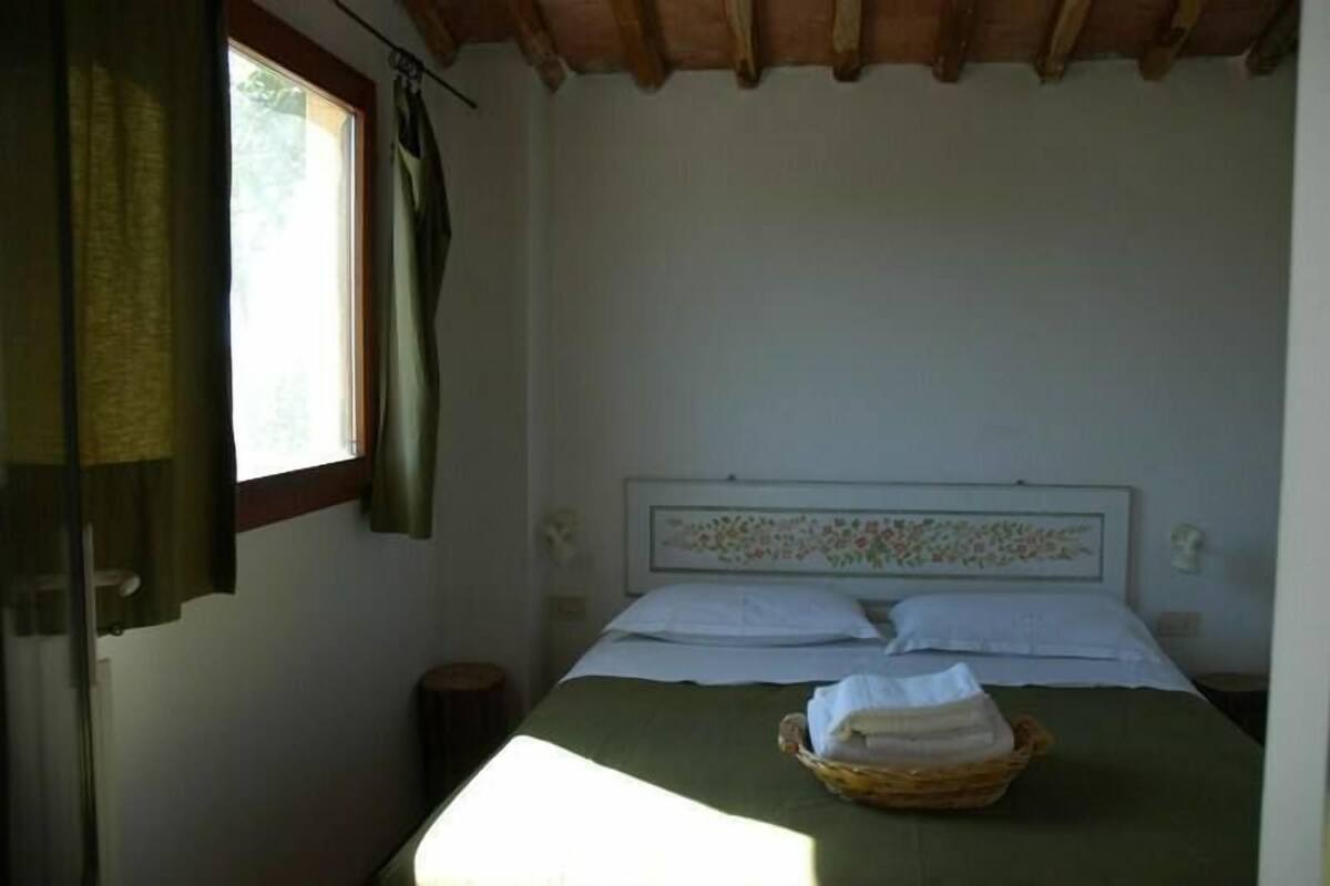 Una cama individual con colcha verde, cabecera estampada con motivos florales, una cesta de toallas dobladas y una ventana con cortinas.