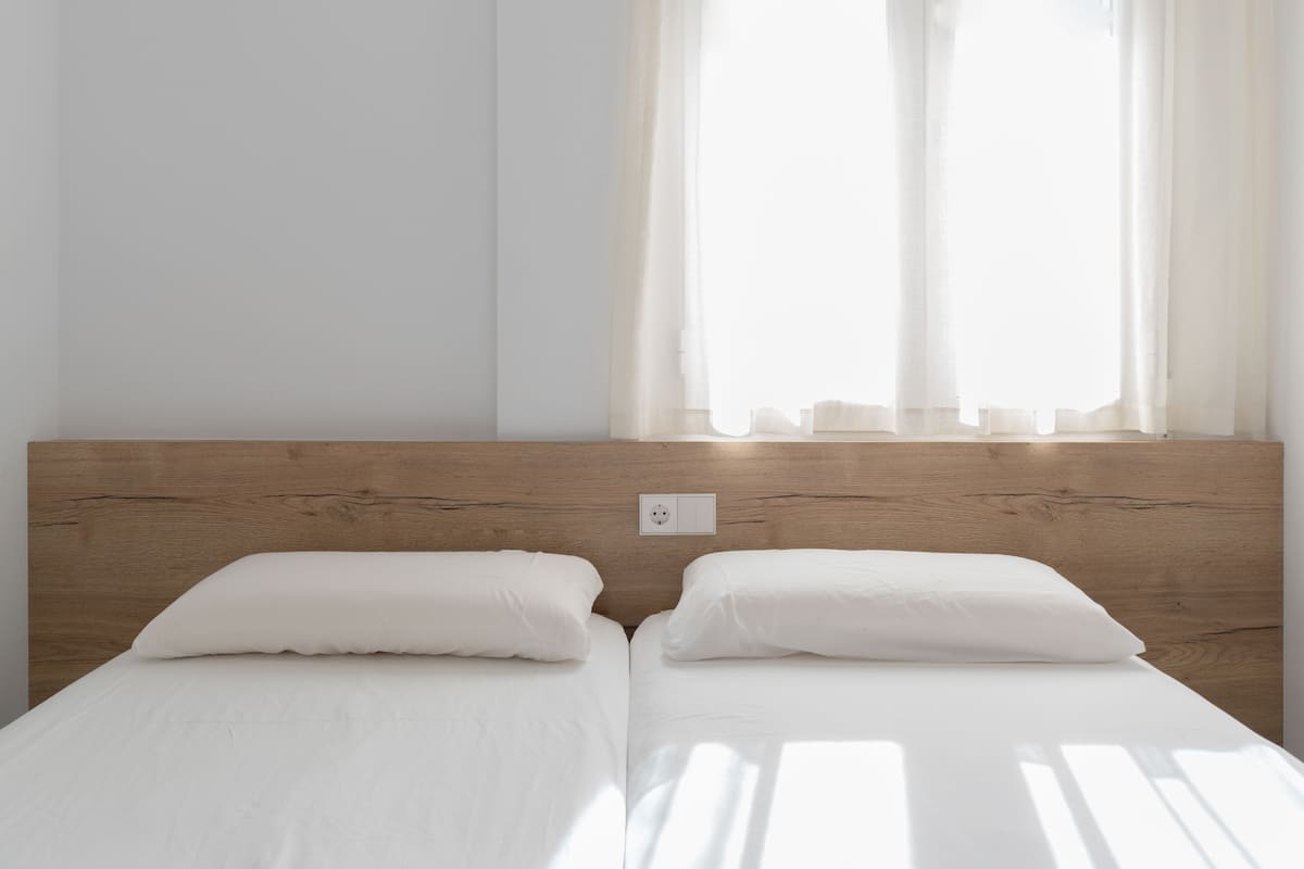 A double bed with white linens in a room with a wooden headboard and a window with sheer curtains.