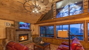 A cozy cabin interior with a stone fireplace, leather seating, a television, and large windows offering a mountain view. - Mountain Views | 4 King Bedroom Luxury Cabin | Hot Tub + Fire Pit (Blue Ridge)