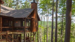 A wooden cabin with a balcony, surrounded by tall trees and a forested landscape. - Mountain Views | 4 King Bedroom Luxury Cabin | Hot Tub + Fire Pit (Blue Ridge)