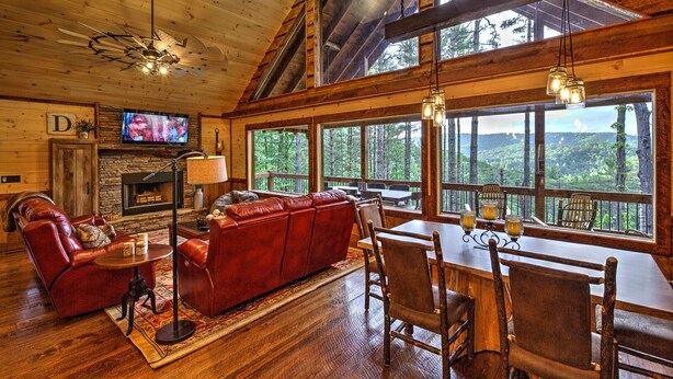 A cozy cabin interior with a fireplace, leather sofa, dining area, and large windows offering a forest view. - Mountain Views | 4 King Bedroom Luxury Cabin | Hot Tub + Fire Pit (Blue Ridge)