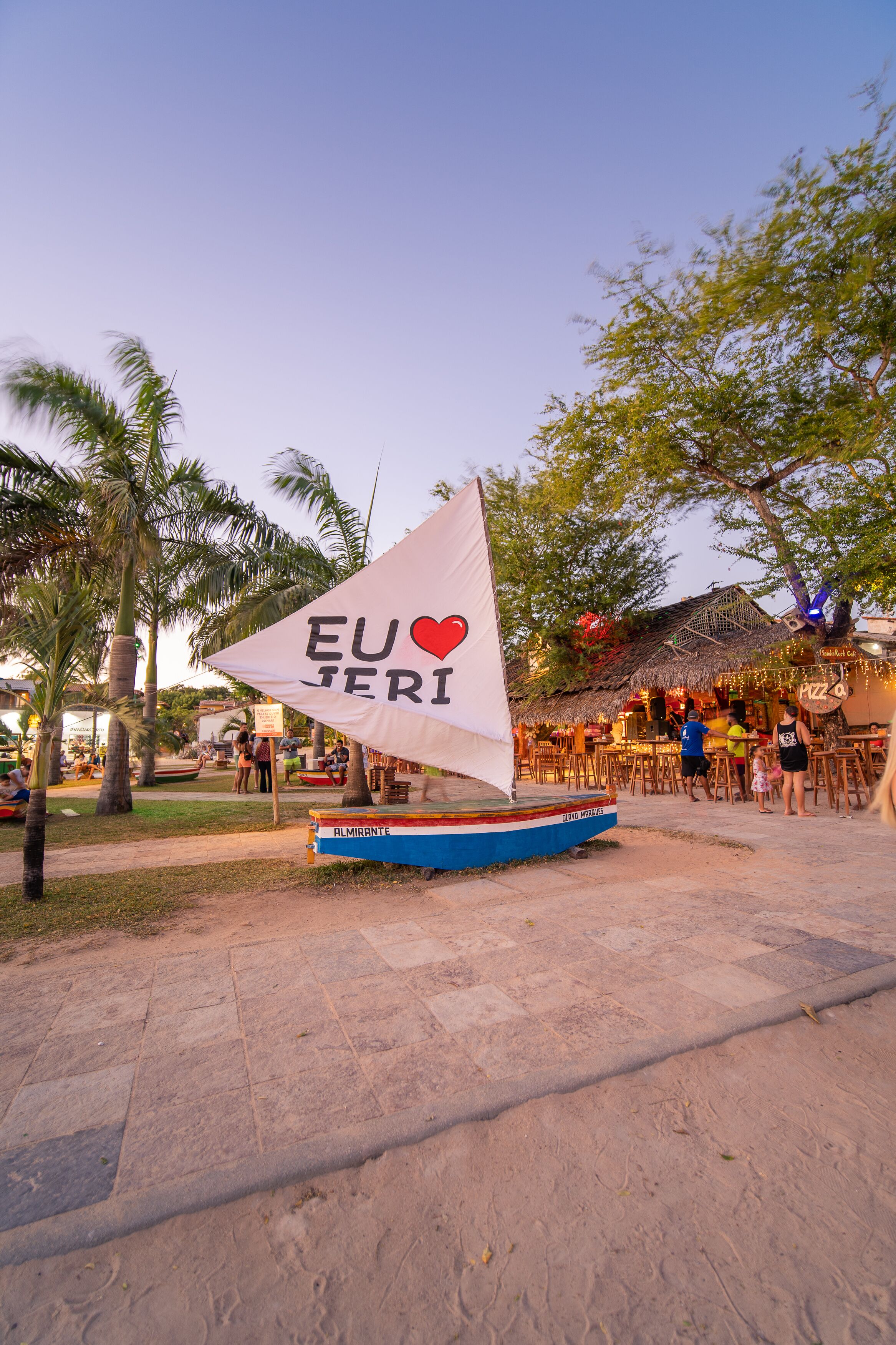 A sailboat with a flag that reads "EU ♥ JERI" in a public area with palm trees and a restaurant in the background.