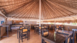 Un restaurante con techo de paja, mesas y sillas de madera, y una cocina visible al fondo.