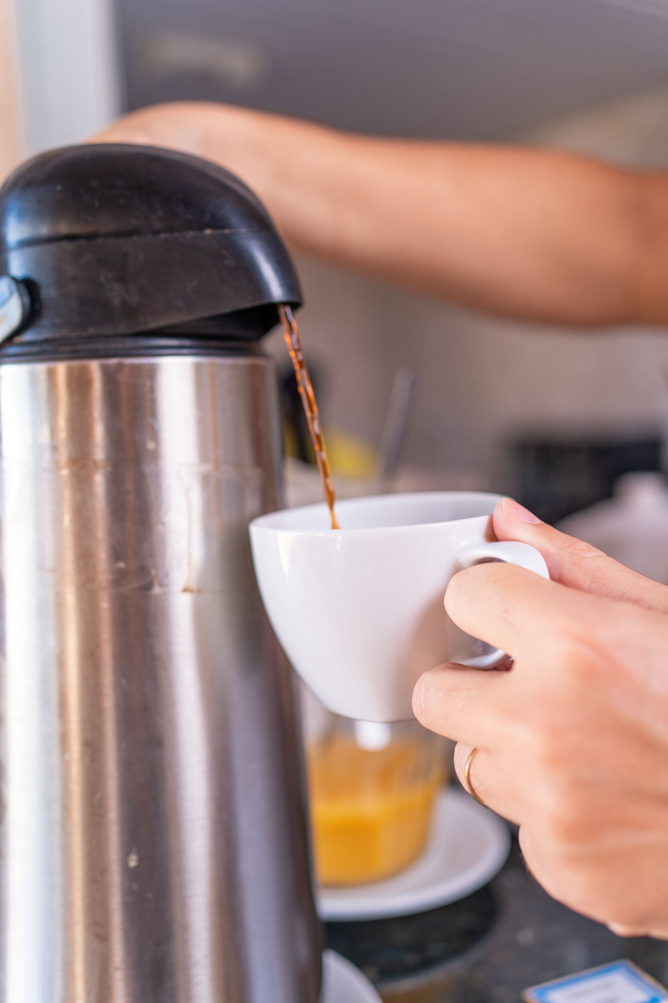 A hand pouring coffee from a stainless steel coffee pot into a white cup.