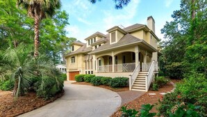 A two-story house with a covered porch, a driveway, and a well-maintained garden with palm trees and shrubs. - 28 Moonshell - Luxury in Folly Field (Hilton Head Island)