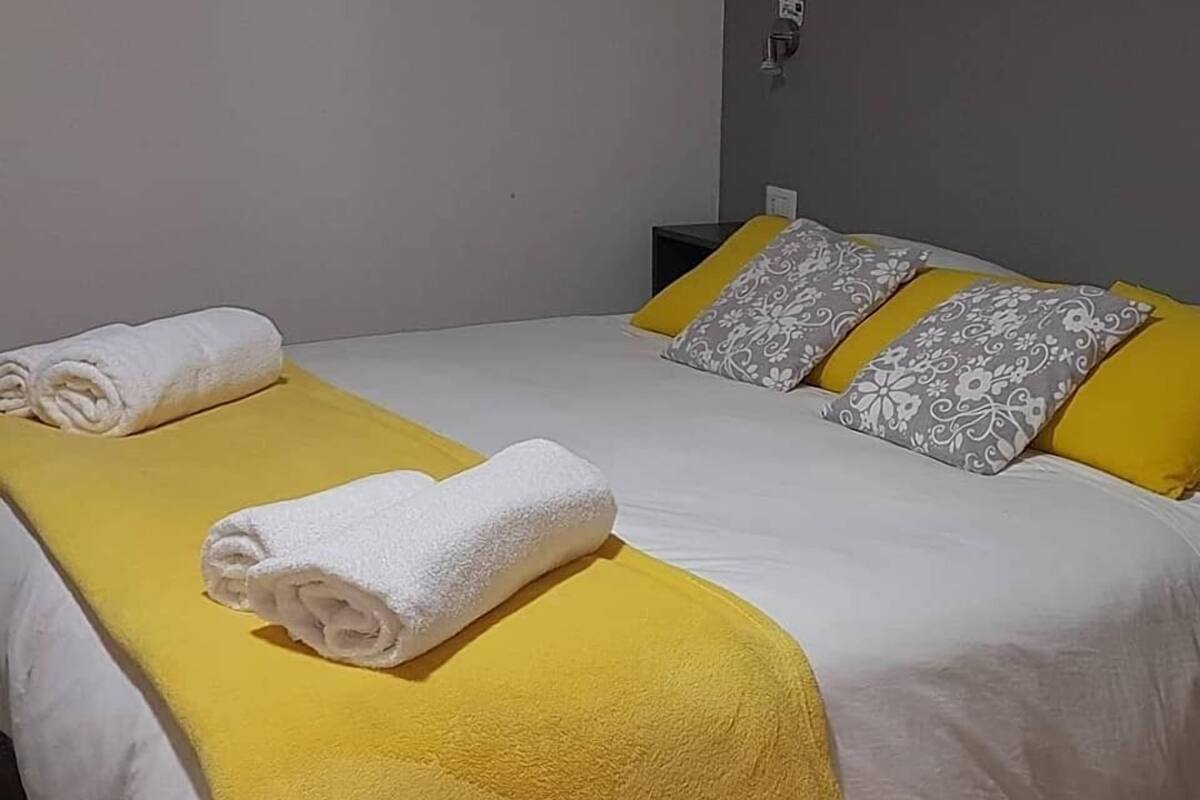 Un lit bien fait, avec une couverture jaune et des oreillers blancs, une petite table de chevet et une lampe fixée au mur.