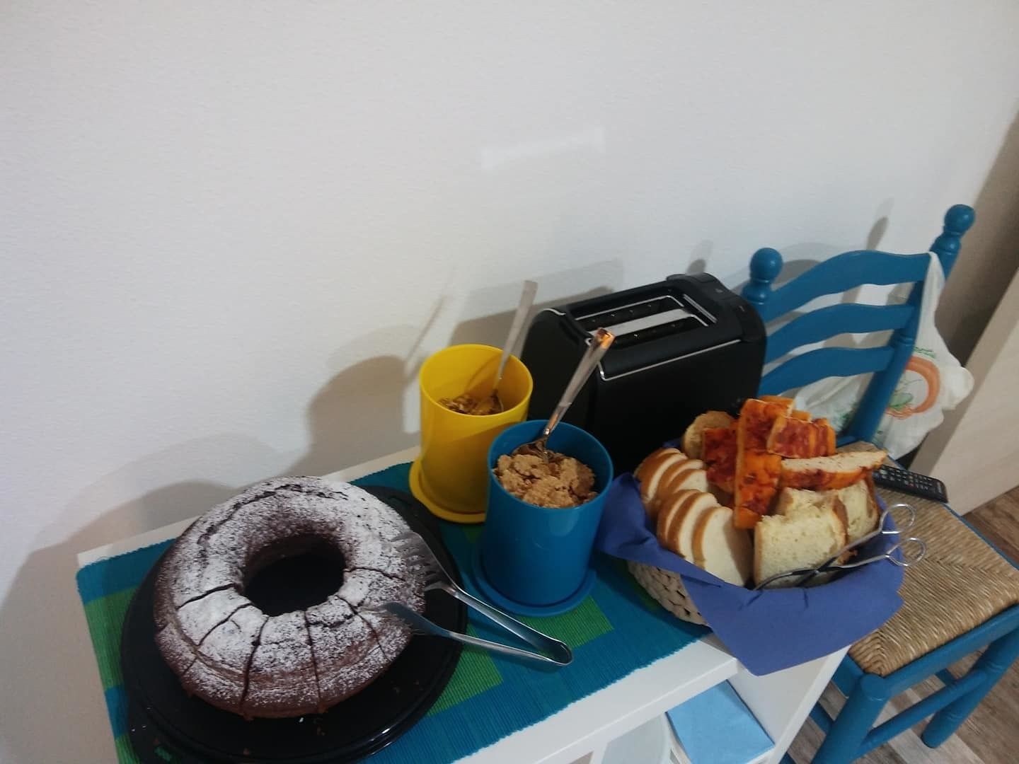 A table with a cake, a toaster, a basket of bread, and a yellow cup.