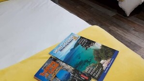 A hotel room with two beds, each with yellow pillows. There is a nightstand with a bottle and a candle. A travel magazine is on the bed. - B&B Fradama' Holiday Dorgali (Dorgali)