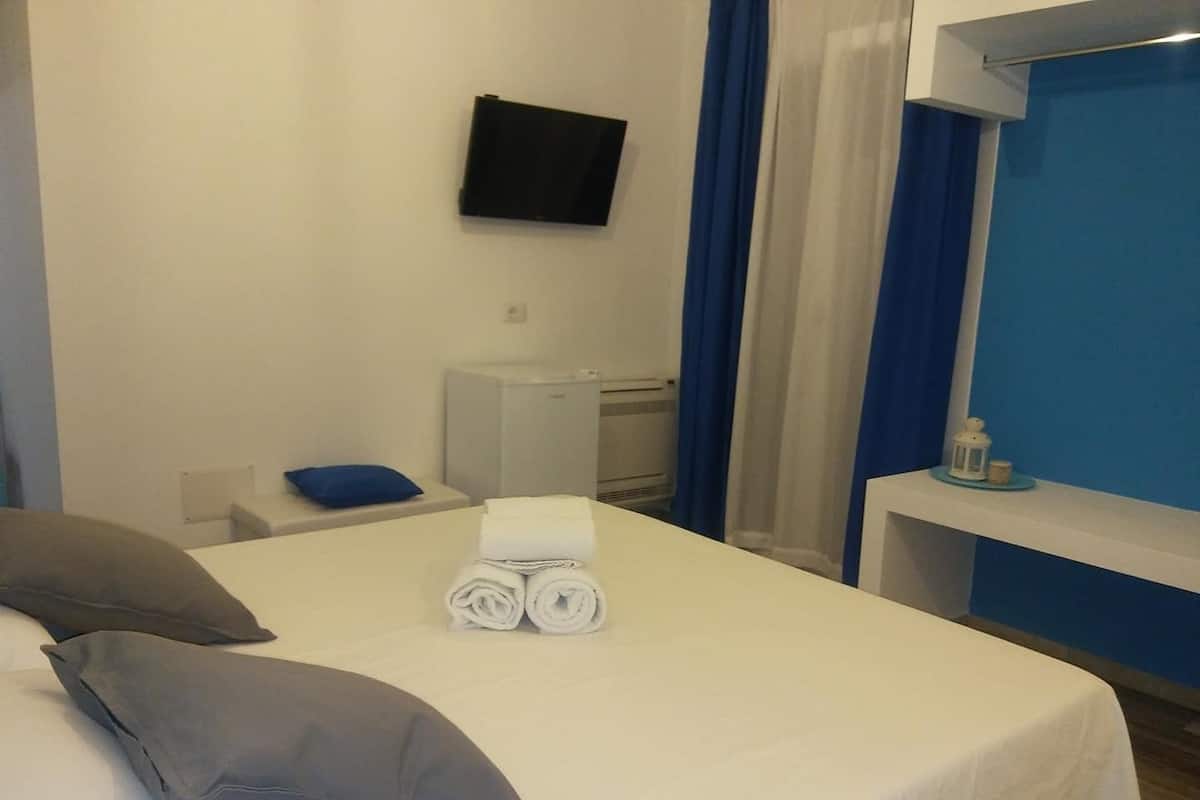 Une chambre d’hôtel avec un lit, un téléviseur fixé au mur, un petit bureau avec une lampe et une fenêtre avec des rideaux.
