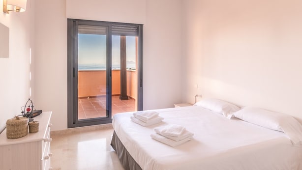 Een hotelkamer met een bed, een nachtkastje, een lamp, uitzicht op zee door een schuifdeur en een kleine tafel met een lamp en decoratieve objecten.