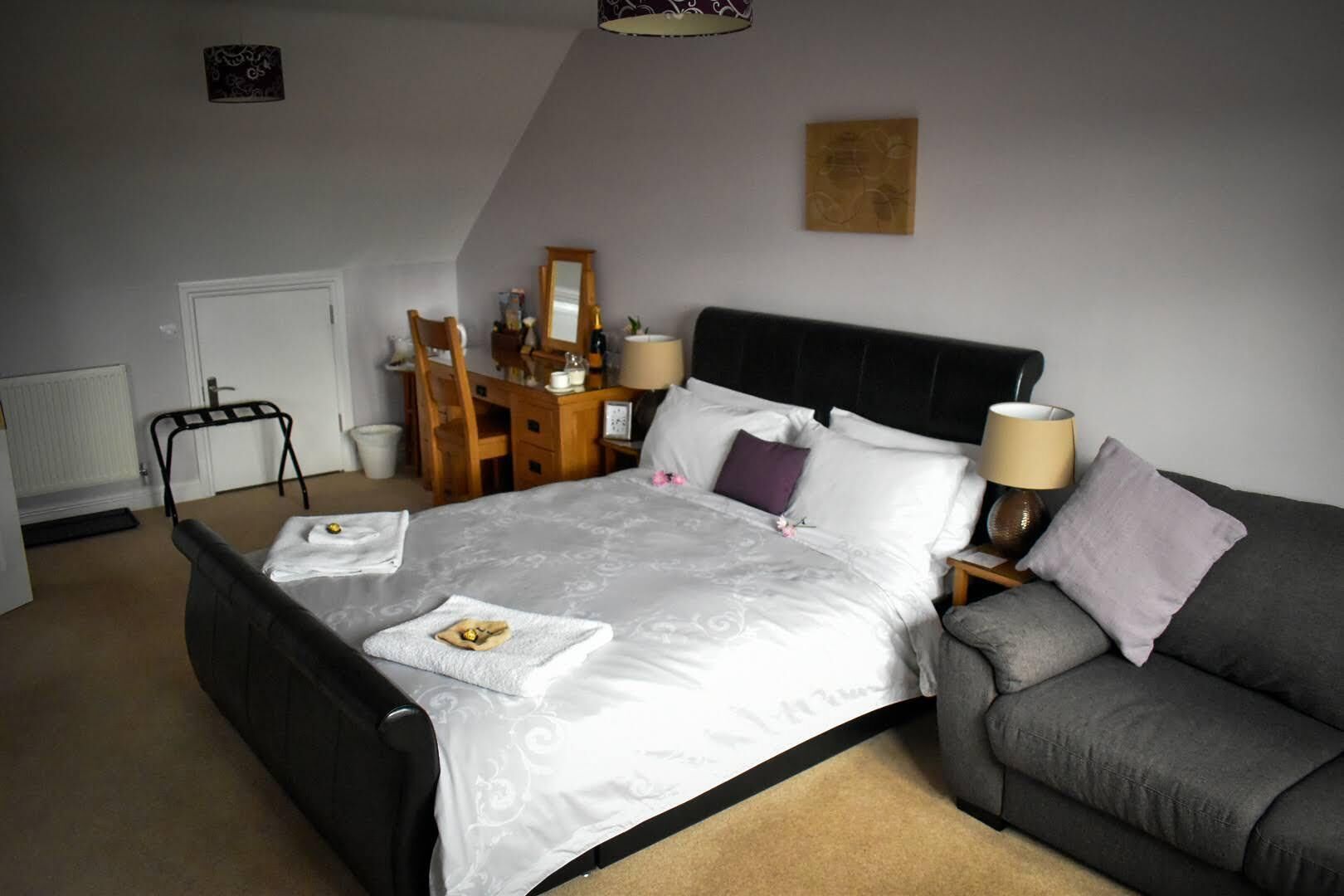 A bedroom with a large bed, a bedside table with lamps, a grey sofa, a wooden desk, and a chair.