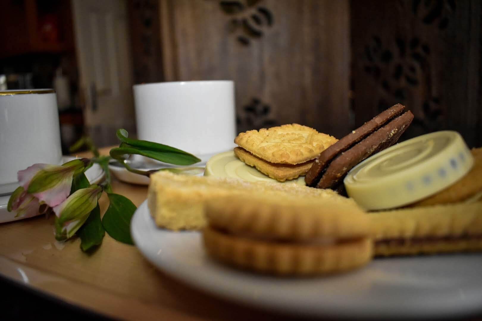 Un plato con varios pasteles y galletas sobre una mesa, junto a una taza blanca y un pequeño jarrón con flores.