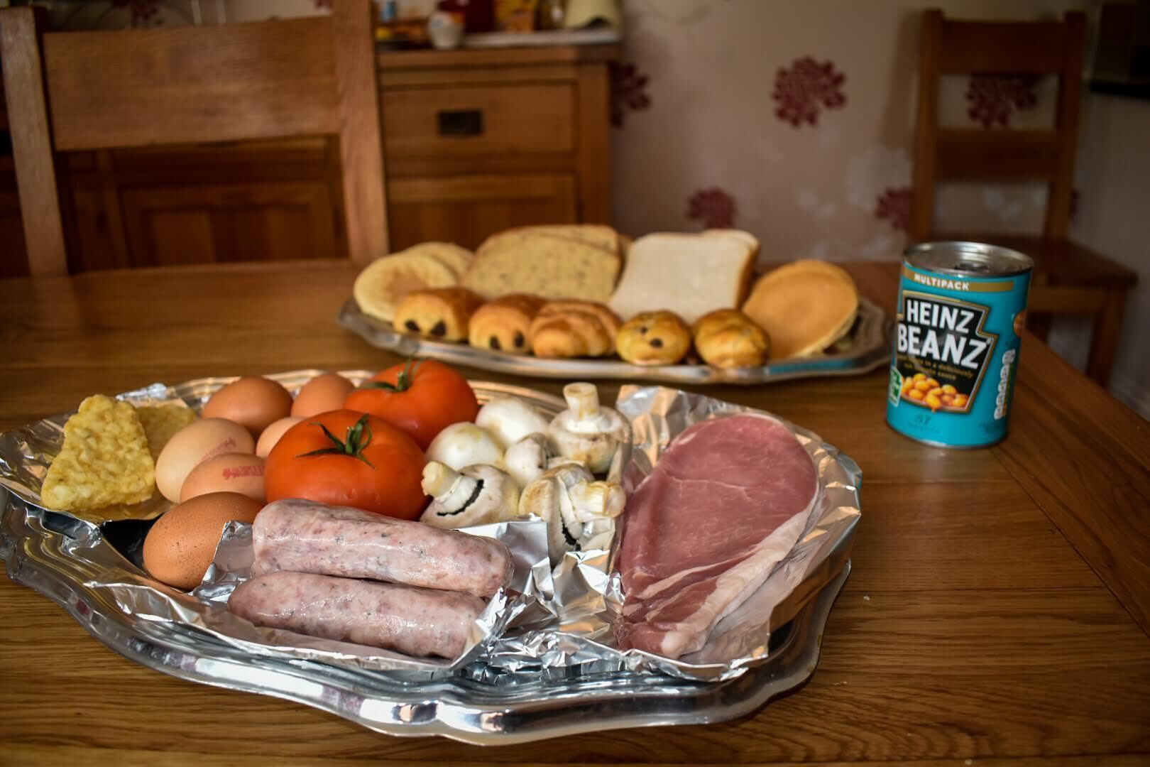A table with food items including sausages, eggs, tomatoes, and mushrooms, and a can of Heinz Beanz.