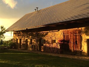 Un bâtiment rustique en pierre, doté d’un toit en pente, de portes en bois et d’une pile de bois de chauffage devant l’entrée.