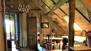 Un intérieur rustique en bois, avec un plafond haut, des poutres apparentes, un coin repas comprenant une table et des chaises, et un lustre.