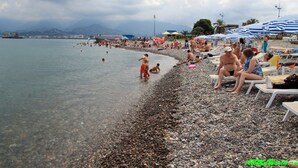 Platja de grava amb gent que pren el sol i nedava, ombrelles i una vista llunyana d'una muntanya.