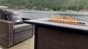 A deck with a fire pit and wicker furniture overlooking a lake with a forested hill in the background.